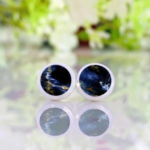 Könnte beinhalten: Ein Paar runde Ohrstecker. Jeder Ohrring hat einen dunkelblauen und goldenen Stein, der in einer weißen Metallfassung gefasst ist. Die Steine haben ein Wirbelmuster. Die Ohrringe werden auf einer reflektierenden Oberfläche präsentiert.