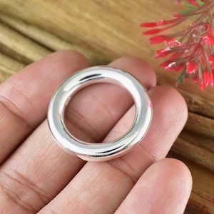 Puede incluir: Un anillo plateado brillante con un diseño liso y redondeado. El anillo es circular con un centro hueco. El fondo incluye una superficie de madera y acentos florales rojos.