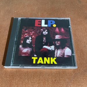 May include: A CD case with a black background and a colourful image of a band called "ELP." The band members are wearing casual clothing and are standing in front of a red and green background. The text "TANK" is printed in yellow letters below the band's name.