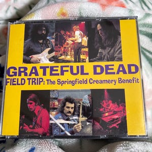 May include: A CD case with a yellow border and the words "Grateful Dead" in purple. The CD cover features band members performing live, with the text "FIELD TRIP: The Springfield Creamery Benefit".