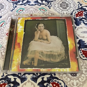 May include: A CD case featuring a vintage image of a woman in a white dress, with the text "Disc Two". The image has a warm, orange and yellow border. The case is on a patterned fabric.