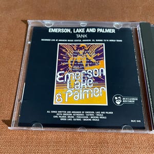 May include: A CD case with a black background and a yellow, orange, and red graphic of the band Emerson, Lake & Palmer. The text "Tank" is printed in white above the graphic. The text "Recorded Live at Anaheim Music Center, Anaheim, CA, during 73/74 World Tours" is printed in white below the graphic. The text "All songs written and arranged by Emerson, Lake and Palmer" is printed in white below the graphic. The text "Keith Emerson: Keyboards/Guitars/Vocals" is printed in white below the graphic. The text "Carl Palmer: Drums/Percussion/Backing Vocals" is printed in white below the graphic. The text "Gregg Lake: Guitars/Backing Vocals" is printed in white below the graphic. The text "Buccaneer Records" is printed in white below the graphic. The text "BUC 046" is printed in white below the graphic.