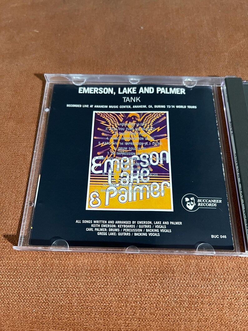 May include: A CD case with a black background and a yellow, orange, and red graphic of the band Emerson, Lake & Palmer. The text "Tank" is printed in white above the graphic. The text "Recorded Live at Anaheim Music Center, Anaheim, CA, during 73/74 World Tours" is printed in white below the graphic. The text "All songs written and arranged by Emerson, Lake and Palmer" is printed in white below the graphic. The text "Keith Emerson: Keyboards/Guitars/Vocals" is printed in white below the graphic. The text "Carl Palmer: Drums/Percussion/Backing Vocals" is printed in white below the graphic. The text "Gregg Lake: Guitars/Backing Vocals" is printed in white below the graphic. The text "Buccaneer Records" is printed in white below the graphic. The text "BUC 046" is printed in white below the graphic.