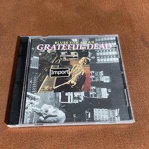 May include: A compact disc case featuring the album "Blues for Allah" by the Grateful Dead. The cover art includes a skeleton, architectural elements, and the word "Import". The case is black with a clear plastic front.