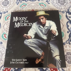 May include: Album cover for "Mixin' Up the Medicine" featuring a seated man in a white shirt and hat, holding a guitar. The album title is in large white letters. Below the title is the text "The Safety Tape June-October 1967".