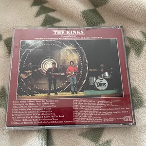 May include: A CD compilation of The Kinks, featuring a band photo on the cover. The album title and tracklist are visible. The CD case is in a clear plastic sleeve, and the background is a patterned fabric.