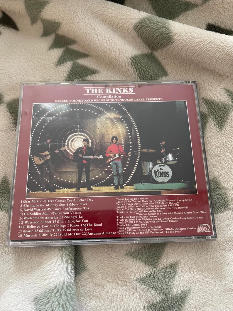 May include: A CD compilation of The Kinks, featuring a band photo on the cover. The album title and tracklist are visible. The CD case is in a clear plastic sleeve, and the background is a patterned fabric.