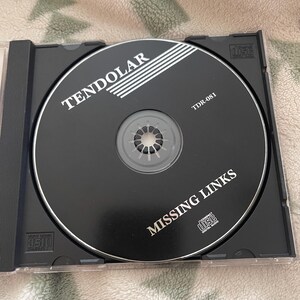 May include: A black compact disc with the text "TENDOLAR" and "MISSING LINKS" in white. The disc is in a clear plastic case. The disc also has the text "TDR-081".