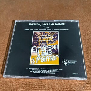 May include: A black CD case with a white and yellow image of Emerson, Lake & Palmer. The text "Tank" is printed in white on a black background. The text "Recorded Live at Anaheim Music Center, Anaheim, CA, during 73/74 World Tours" is printed in white on a black background. The text "All songs written and arranged by Emerson, Lake and Palmer" is printed in white on a black background. The text "Keith Emerson: Keyboards/Guitars/Vocals" is printed in white on a black background. The text "Carl Palmer: Drums/Percussion/Backing Vocals" is printed in white on a black background. The text "Gregg Lake: Guitars/Backing Vocals" is printed in white on a black background. The text "Buccaneer Records" is printed in white on a black background. The text "BUC 046" is printed in white on a black background.