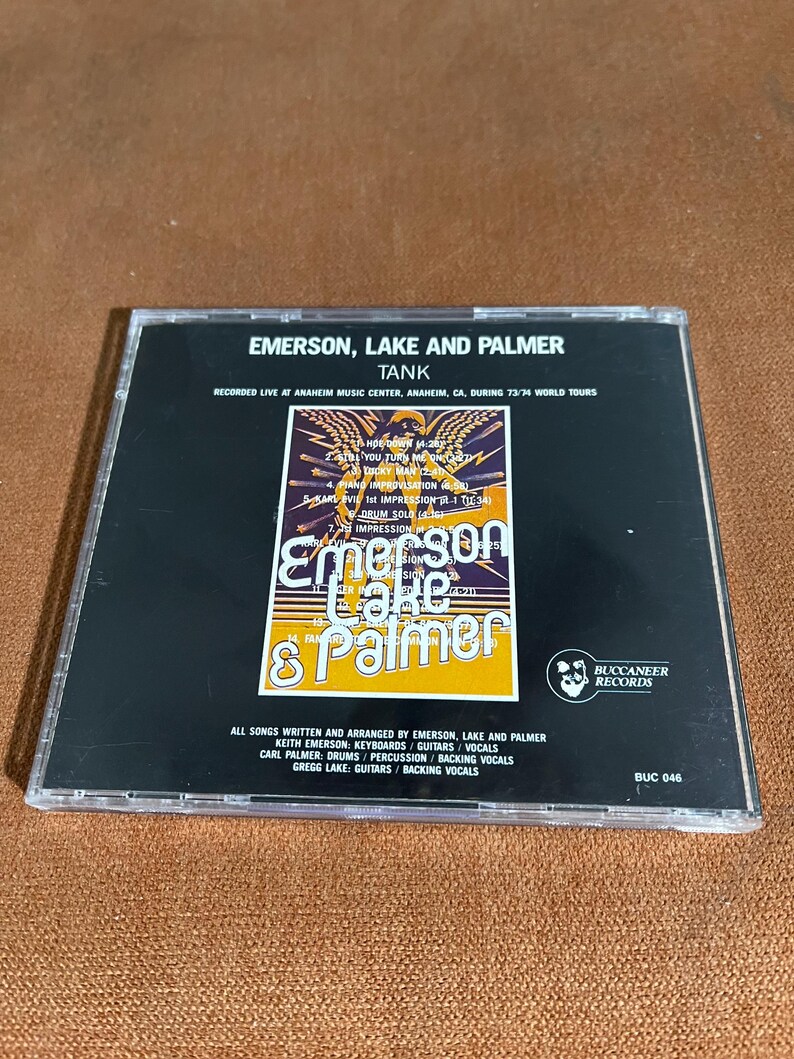 May include: A black CD case with a white and yellow image of Emerson, Lake & Palmer. The text "Tank" is printed in white on a black background. The text "Recorded Live at Anaheim Music Center, Anaheim, CA, during 73/74 World Tours" is printed in white on a black background. The text "All songs written and arranged by Emerson, Lake and Palmer" is printed in white on a black background. The text "Keith Emerson: Keyboards/Guitars/Vocals" is printed in white on a black background. The text "Carl Palmer: Drums/Percussion/Backing Vocals" is printed in white on a black background. The text "Gregg Lake: Guitars/Backing Vocals" is printed in white on a black background. The text "Buccaneer Records" is printed in white on a black background. The text "BUC 046" is printed in white on a black background.