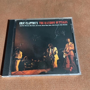 May include: A CD case featuring Eric Clapton's "The Rainbow Outtakes." The cover displays a live performance photo of Clapton and his band on stage. The text includes the album title and the names of the musicians.