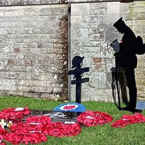 May include: A black metal silhouette of a soldier standing to attention in front of a stone wall. The soldier is holding a rifle and wearing a helmet. There are red poppy wreaths on the ground in front of the soldier.