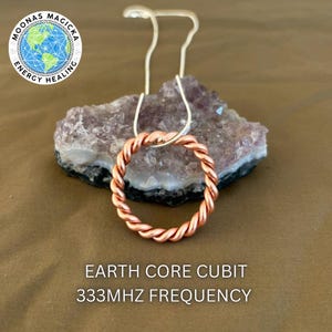 May include: A copper-colored, twisted-wire pendant necklace with a circular design. The pendant hangs from a silver-colored chain and rests on a purple crystal. The image includes the text "EARTH CORE CUBIT 333MHZ FREQUENCY".