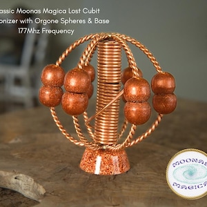 Puede incluir: Un armonizador de metal de color cobre con una base y una columna central con un diseño en espiral. El armonizador tiene seis esferas de color cobre unidas a la columna central. El armonizador está sobre una superficie de madera. El texto "Classic Moonas Magica Lost Cubit Harmonizer with Orgone Spheres & Base 177Mhz Frequency" está por encima del armonizador. El texto "MOONAS MAGIC" está por debajo del armonizador.