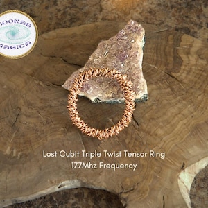 May include: A copper-colored Lost Cubit Triple Twist Tensor Ring with a 177Mhz frequency. The ring is on a wooden surface with a piece of pink and white crystal.
