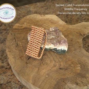 Op de afbeelding: Een koperen spoel op een houten oppervlak met een paarse en witte kristal. De spoel is gelabeld "Sacred Cubit Transmutation Coil 144Mhz Frequency Transmutes density into Light".