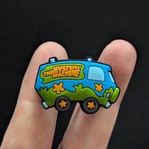 May include: A blue and green shoe charm shaped like the Mystery Machine van from Scooby-Doo. The van has the words "The Mystery Machine" written on the side.