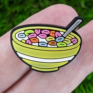 May include: A yellow bowl with a white rim filled with colorful cereal and a white spoon. The bowl is sitting on a finger.