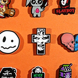 May include: A collection of colorful shoe charms with various designs. Includes a cross with a face, a smiley face, a teddy bear with a knife, a bunny with a knife, and a skull heart. The word "SLAYBOY" is also visible.