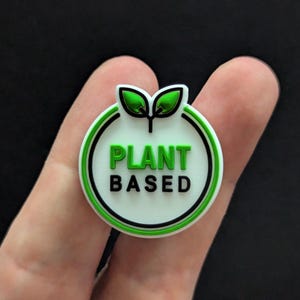 May include: A white circle with a green outline and two green leaves at the top. The circle has the text "PLANT BASED" in black letters.