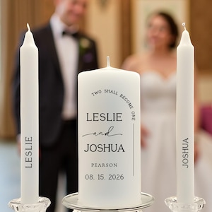 May include: A white unity candle set for a wedding ceremony. The set includes a large pillar candle with the text "TWO SHALL BECOME ONE" and the names "LESLIE and JOSHUA" and two taper candles with the names "LESLIE" and "JOSHUA".