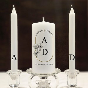 May include: Two white unity candles on glass candle holders. The center candle has a gold frame with the text "Angelina & Dominic A + D November 25, 2023" and a floral design.