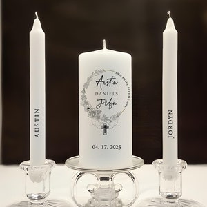 May include: A set of three white candles on clear glass holders. The two side taper candles are printed with the names Austin and Jordyn. The center pillar candle features a floral wreath, a cross, the text Austin, Daniels, Jordyn, Two Shall Become One, and the date 04. 17. 2025.