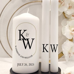 May include: A white unity candle set with two taper candles. The large candle has the initials "K & W" and the names "Katherine and William" with the date "July 26, 2023". The taper candles have the initials "KW". The candles are on a black base.