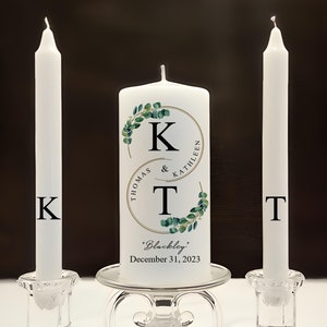 May include: Two white unity candles with the initials "K" and "T" on them. The center candle has a gold circle with the initials "K" and "T" inside, surrounded by a green eucalyptus wreath. The text "Thomas & Kathleen" is written around the circle. The text "Blackley" and "December 31, 2023" is written below the circle.