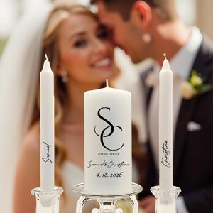 May include: A white unity candle set for a wedding ceremony. The set includes a large pillar candle with the initials "SC" and the names Samuel & Christine, and two taper candles with names. The date 4.18.2026 is also printed on the pillar candle.