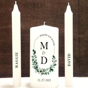 May include: Three white wedding unity candles with the names Maggie and David printed on them. The center candle has a gold and green eucalyptus wreath with the initials M & D and the date 11.27.2023.