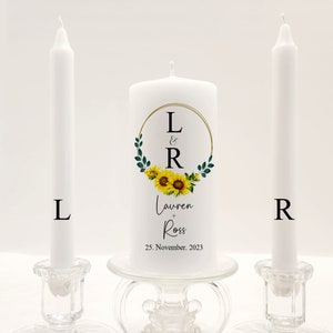 May include: Two white unity candles with a single larger candle in the center. The center candle has a sunflower wreath design with the initials L & R and the names Lauren + Ross and the date 25. November 2023. The candles are on glass candle holders.
