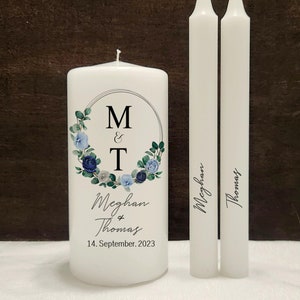 May include: Two white pillar candles with the names "Meghan" and "Thomas" written on them. A larger white candle with a floral wreath design and the initials "M&T" in a circle, the names "Meghan & Thomas" and the date "14. September. 2023".