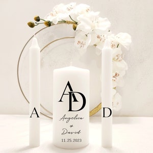 May include: Two white pillar candles with the initials "A" and "D" on them. The center candle has the initials "A" and "D" intertwined and the text "Angelica & David 11.25.2023".