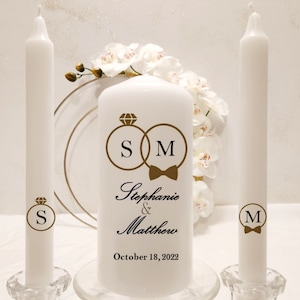 May include: Three white unity candles with gold rings and initials "S" and "M" on the sides. The centre candle has the initials "SM" with a bow tie and the text "Stephanie & Matthew October 18, 2022".