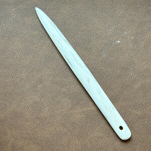 May include: A light-coloured bone folder tool with a pointed end and a small hole at the rounded end. The tool is used for smoothing paper and other materials. The bone folder is approximately 20 cm long.