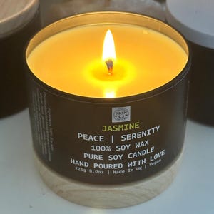 Könnte beinhalten: Ein schwarzes Kerzengefäß mit einem Holzdeckel. Die Kerze ist angezündet und das Wachs ist leuchtend gelb. Das Etikett auf dem Gefäß lautet "Jasmine", "Peace | Serenity", "100% Soy Wax", "Pure Soy Candle", "Hand Poured With Love", "225g 8.0oz", "Made in UK" und "Vegan".