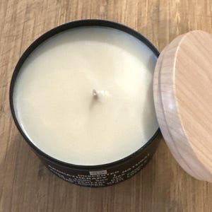 May include: A black candle jar with a wooden lid. The candle is white and unscented. The jar has text on the bottom that reads "UNSCENTED NATURAL THERAPY".