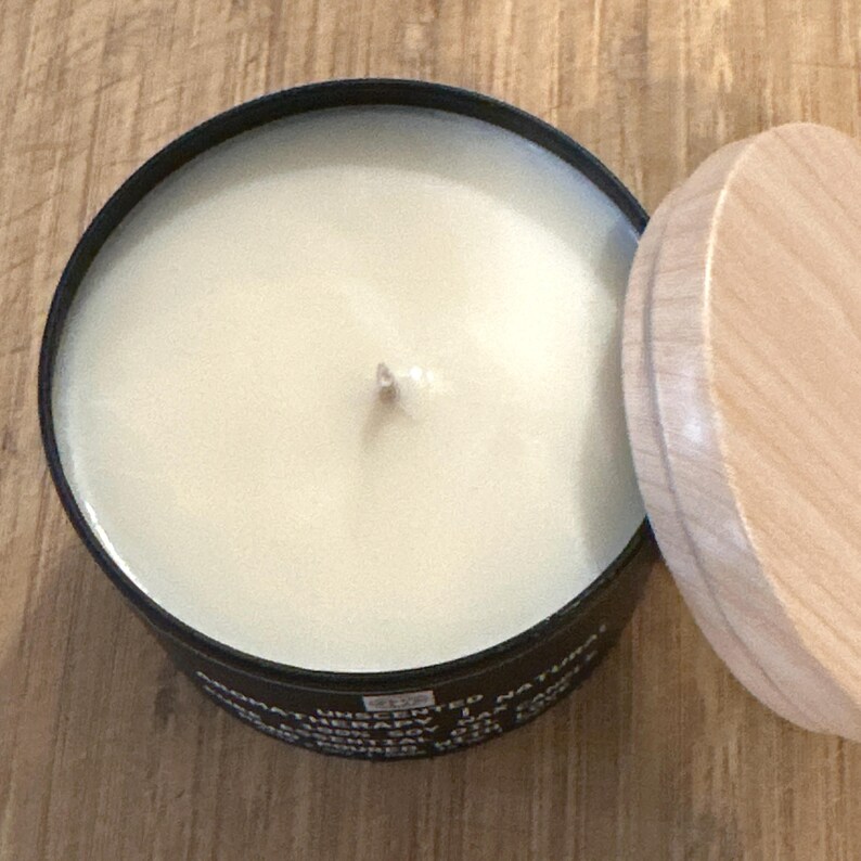 May include: A black candle jar with a wooden lid. The candle is white and unscented. The jar has text on the bottom that reads "UNSCENTED NATURAL THERAPY".