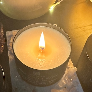 May include: A black candle jar with a white soy wax candle burning inside. The candle is labelled "Handcrafted Natural Soy Wax Candle" and "Made with Love".