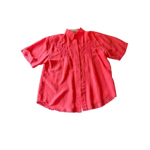 May include: A bright coral-colored short-sleeved button-down shirt. The shirt has embroidered floral detailing on the chest. The fabric appears to be lightweight and suitable for warm weather.