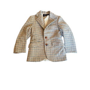 May include: A long-sleeve blazer featuring a brown, tan, and white houndstooth pattern. The garment includes a notched lapel, two front flap pockets, and two brown buttons for closure. The interior is lined with a solid white fabric.