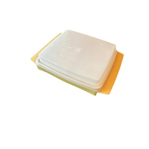 May include: A square, off-white plastic container with a matching lid. The container sits on a yellow and orange base. The lid has a circular design in the center. The container is likely for food storage.