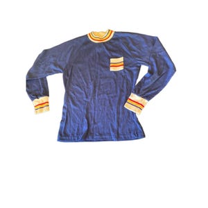 May include: A long-sleeve blue shirt featuring a mock neck collar, white cuffs, and a chest pocket. The collar and cuffs are accented with thin horizontal stripes in red, yellow, and blue. The pocket also displays these same colorful stripes.