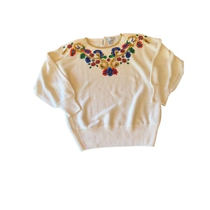 May include: Cream-colored sweater with a crew neck and long sleeves. The neckline and shoulders are adorned with colorful floral embroidery, including red, blue, and gold accents. The sweater has a ribbed hem and cuffs.
