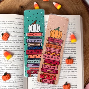 May include: Two decorative bookmarks with a stack of books design, topped with a pumpkin illustration. One bookmark has a teal background with white polka dots, the other has a tan background with small dots. Candy corn and pumpkin-shaped decorations are scattered around.