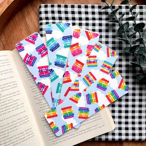 May include: A collection of colorful bookmarks with a repeating pattern of stacked, rainbow-colored shapes against a light blue sky with white clouds. The bookmarks are fanned out over an open book and a black and white checkered surface.