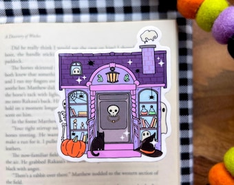May include: A whimsical Halloween-themed sticker featuring a purple house with a skull door knocker, a black cat, and a pumpkin. The sticker also includes a witch's hat, a ghost, and a chimney with smoke.