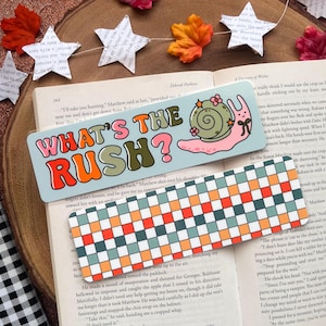 May include: Two colorful bookmarks rest on an open book. One bookmark features the text "WHAT'S THE RUSH?" with a snail illustration. The other has a checkered pattern in orange, green, and blue. Decorative stars and leaves are in the background.