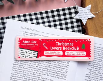 Puede incluir: Un marcapáginas rosa y rojo con el texto "Christmas Lovers Bookclub" y detalles sobre el club de lectura, incluyendo la fecha, la ubicación y las actividades. El marcapáginas presenta un bastón de caramelo, un árbol de Navidad y un libro abierto.